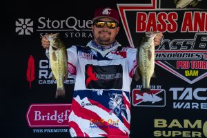 Angler on stage holding two largemouth bass, wearing a red cap and reflective sunglasses, with sponsor banners behind him.