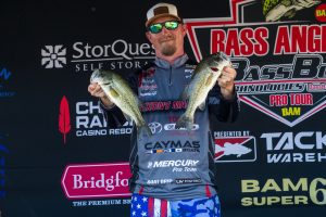 Man in sunglasses and cap holding two largemouth bass on a competition stage with sponsor banners behind him.